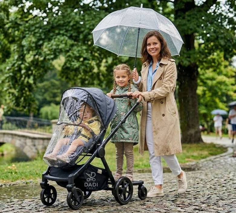 Mama z dwójką dzieci w parku w deszczu, starsze dziecko jedzie na dostawce do wózka spacerowego.